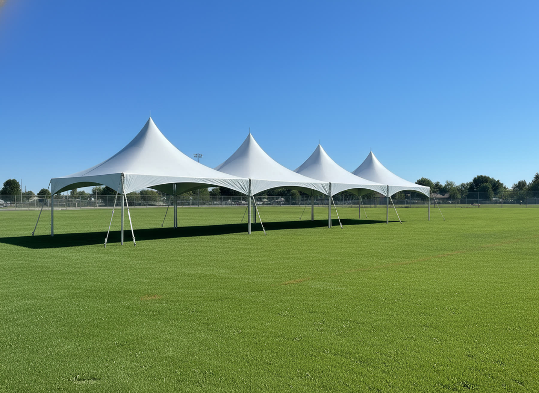 Four white marquee tents in a row on a grass field, three-quarter angle, blue sky overhead