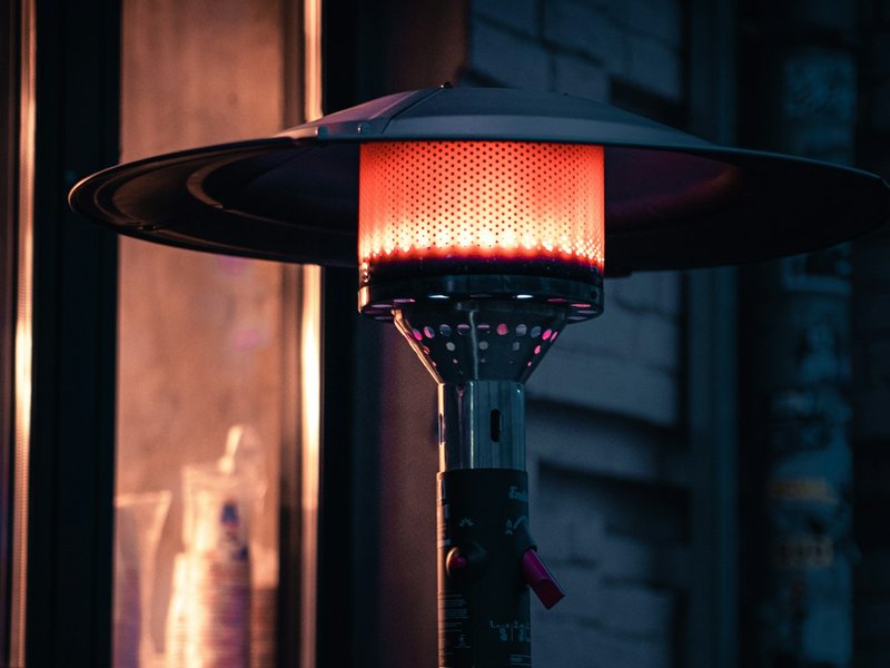 Tall stainless-steel mushroom-hood propane patio heater glowing red against a brick exterior at dusk